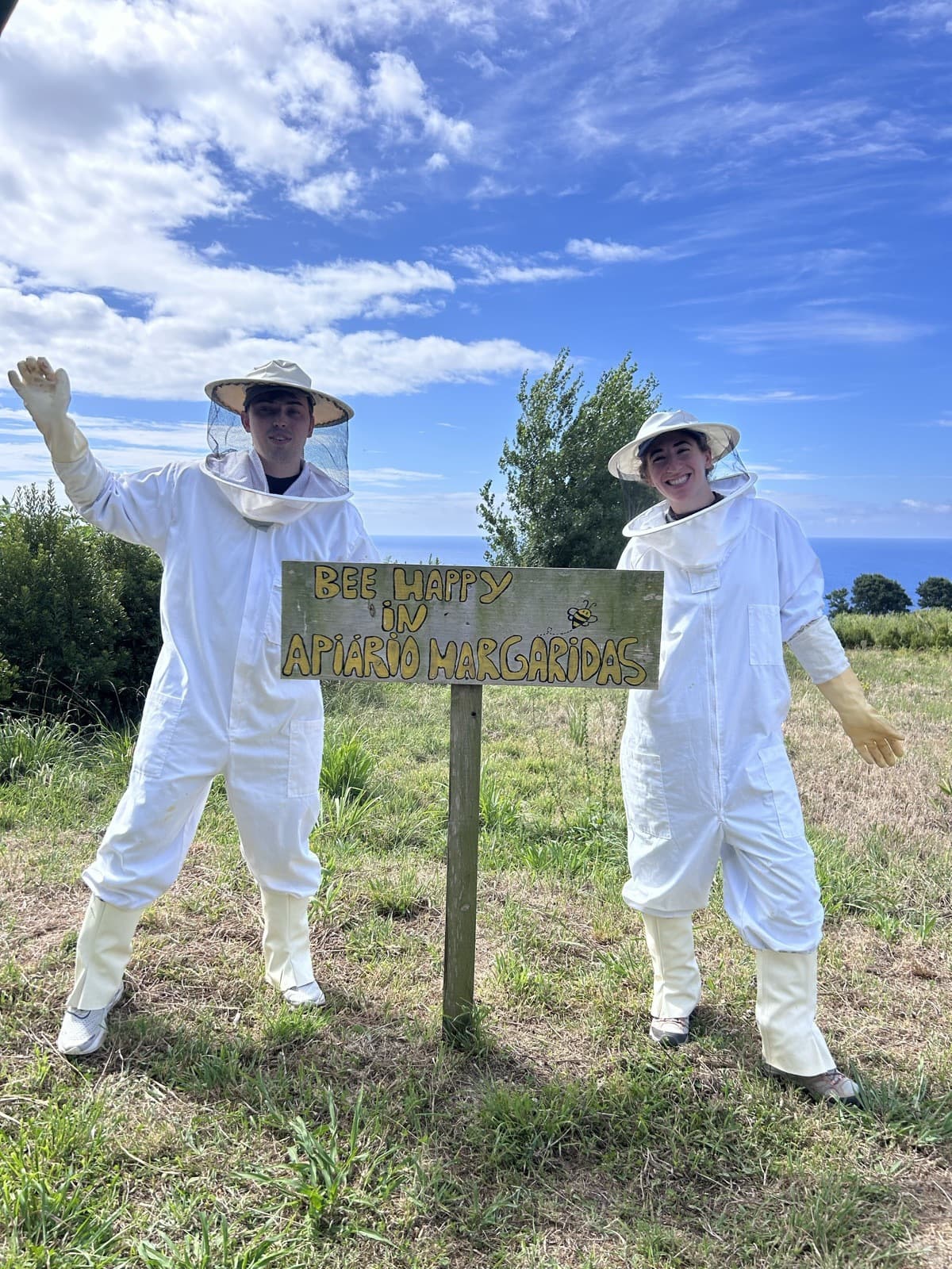 Jen and Luke in beekeeping suits beside an apiary sign